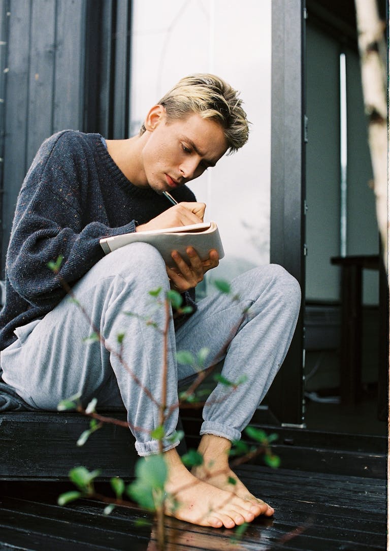 A young man sitting barefoot, writing in a notebook on a wooden deck.