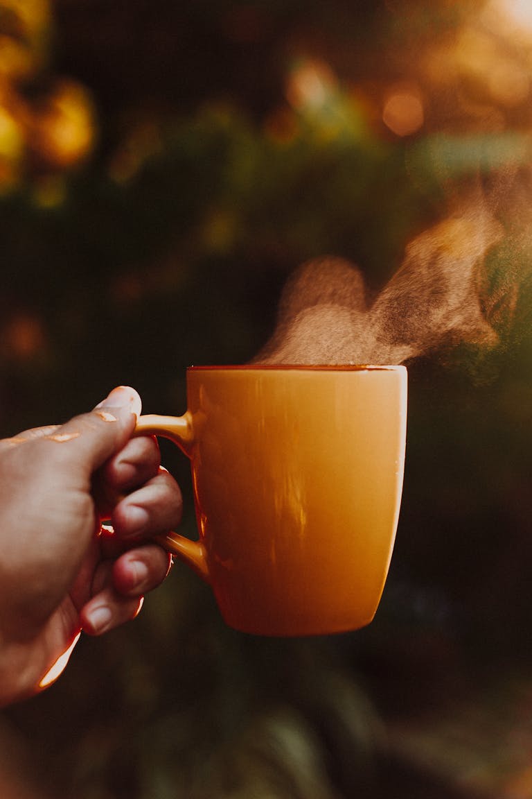 Hand holding a steaming orange mug outdoors in warm morning sunlight, creating a cozy atmosphere.